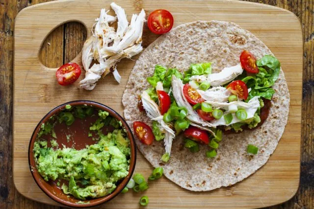 Whole-wheat tortilla topped with shredded chicken, lettuce, tomatoes and scallions on a cutting board, with guacamole bowl nearby.