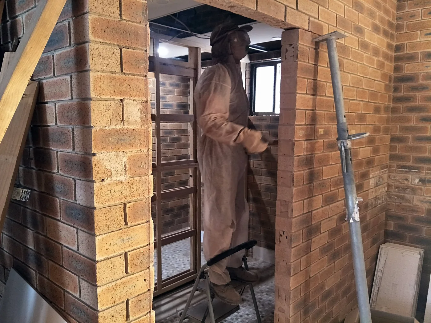 Worker in tan coveralls stands on a step stool inside a brick-walled construction site, fitting a doorway frame among scaffolding.