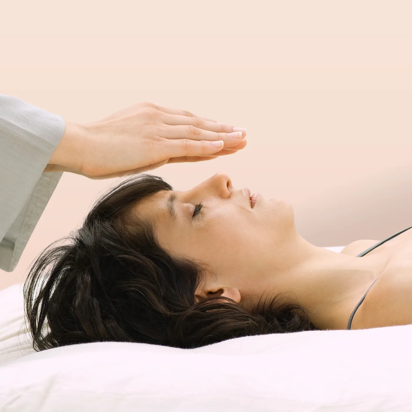 Woman lies in bed with eyes closed while a hand hovers over her forehead, transmitting internal energy like Reiki Healing.