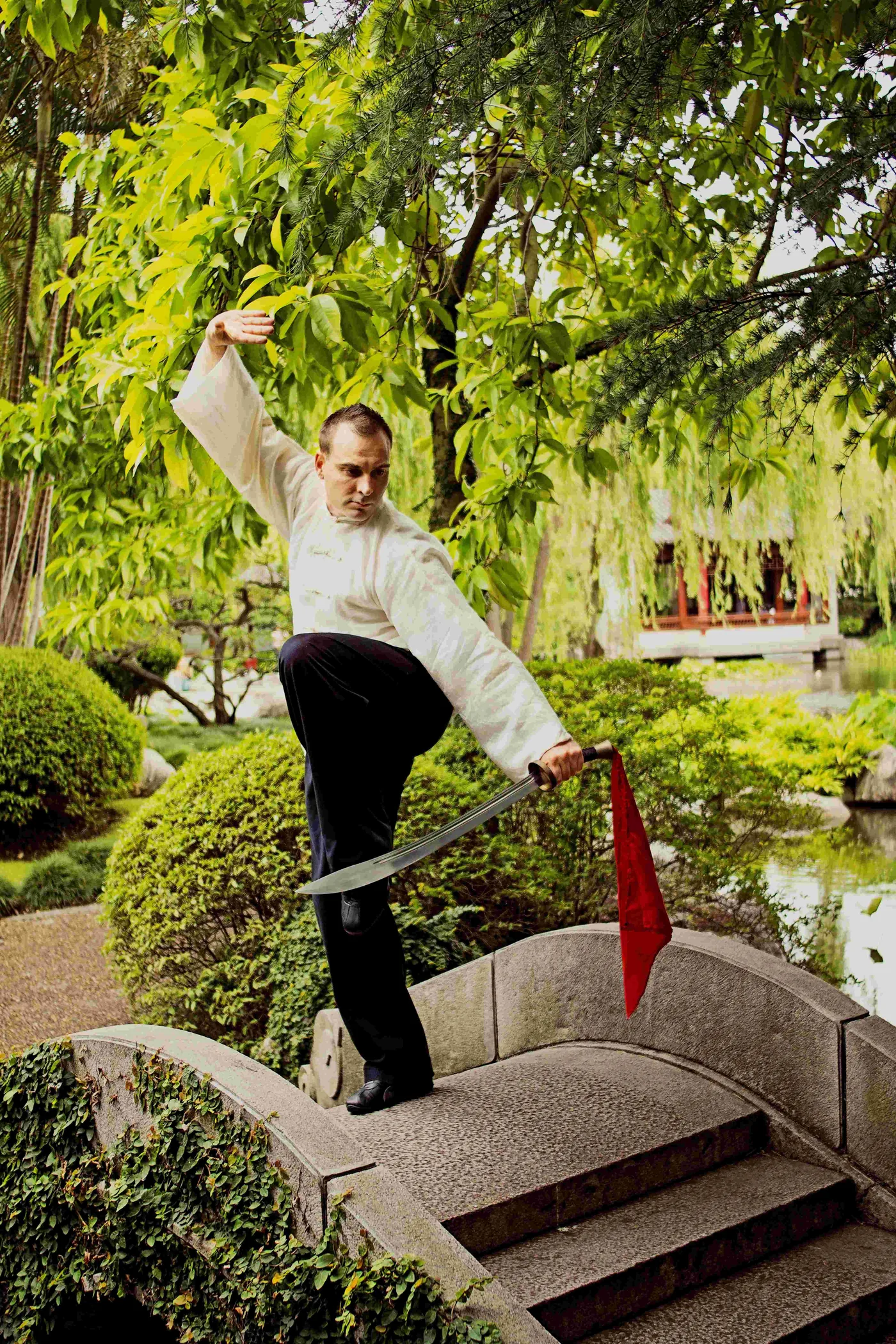 Martial artist in white performs a sword pose on a stone bridge in a lush green garden, red tassel hanging from the blade.