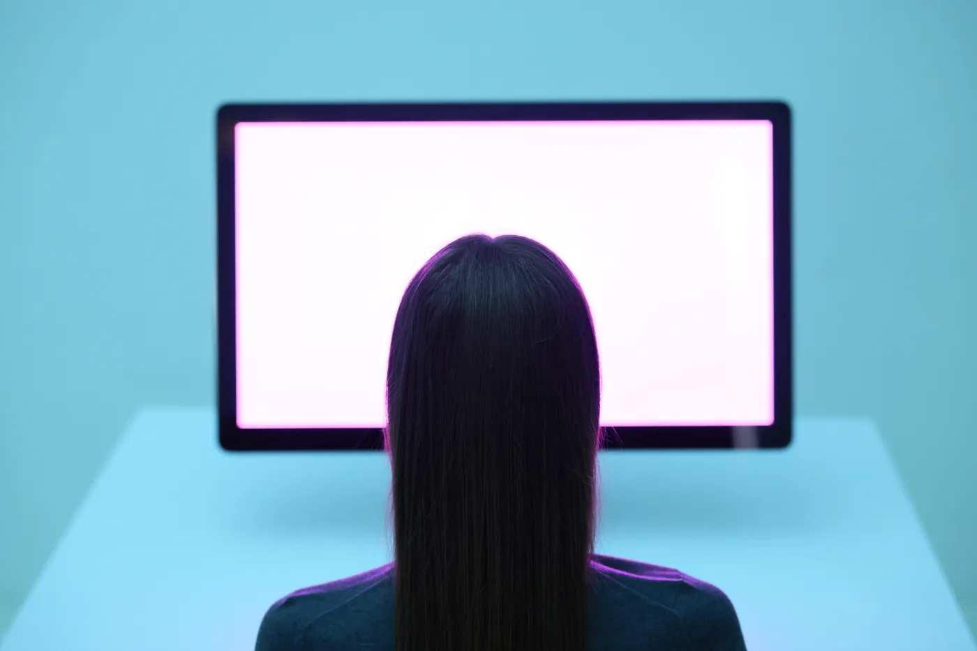 Back view of a person with long dark hair facing a glowing blank monitor in a blue room, with a calm, futuristic mood
