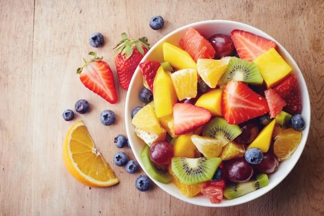 Colorful bowl of mixed fruit—strawberries, kiwi, mango, oranges and blueberries—on a wooden table.