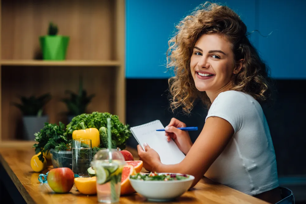 Smiling woman writes in a notebook at a kitchen table with fresh fruit, vegetables and salad on a wooden table.