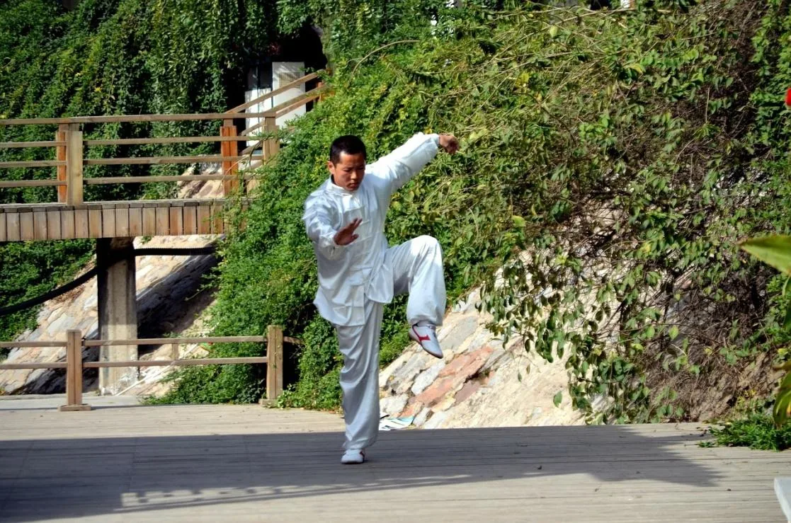 Man in white tai chi outfit balances in a martial arts pose on a wooden deck, with lush greenery and a bridge behind him.