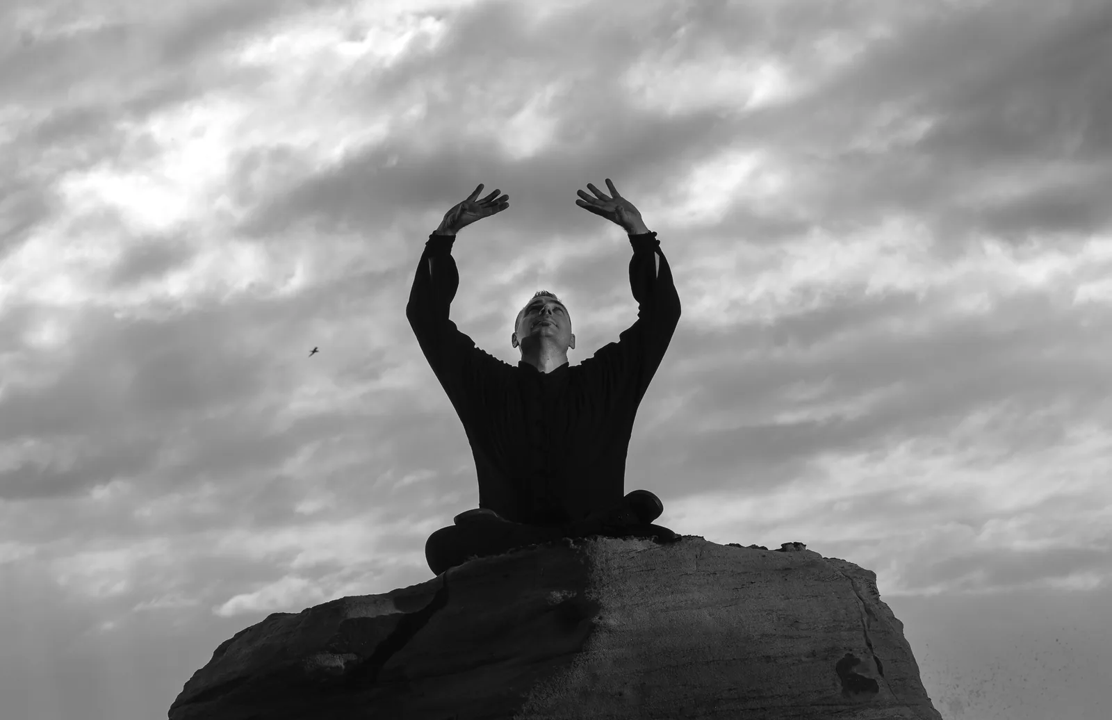 Seated meditation on a sea rock with arms raised to the sky, in black and white.