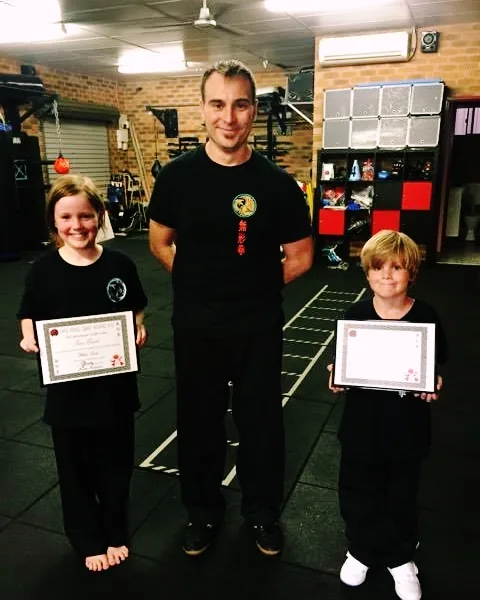 Two young students hold their white sash certificates with their instructor.