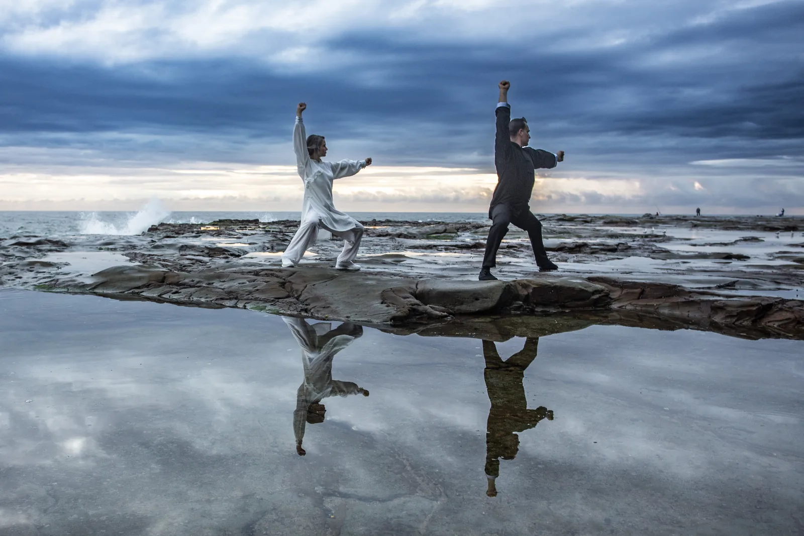 Two Tai Chi practitioners on a sea rock platform, reflected in a rock pool.