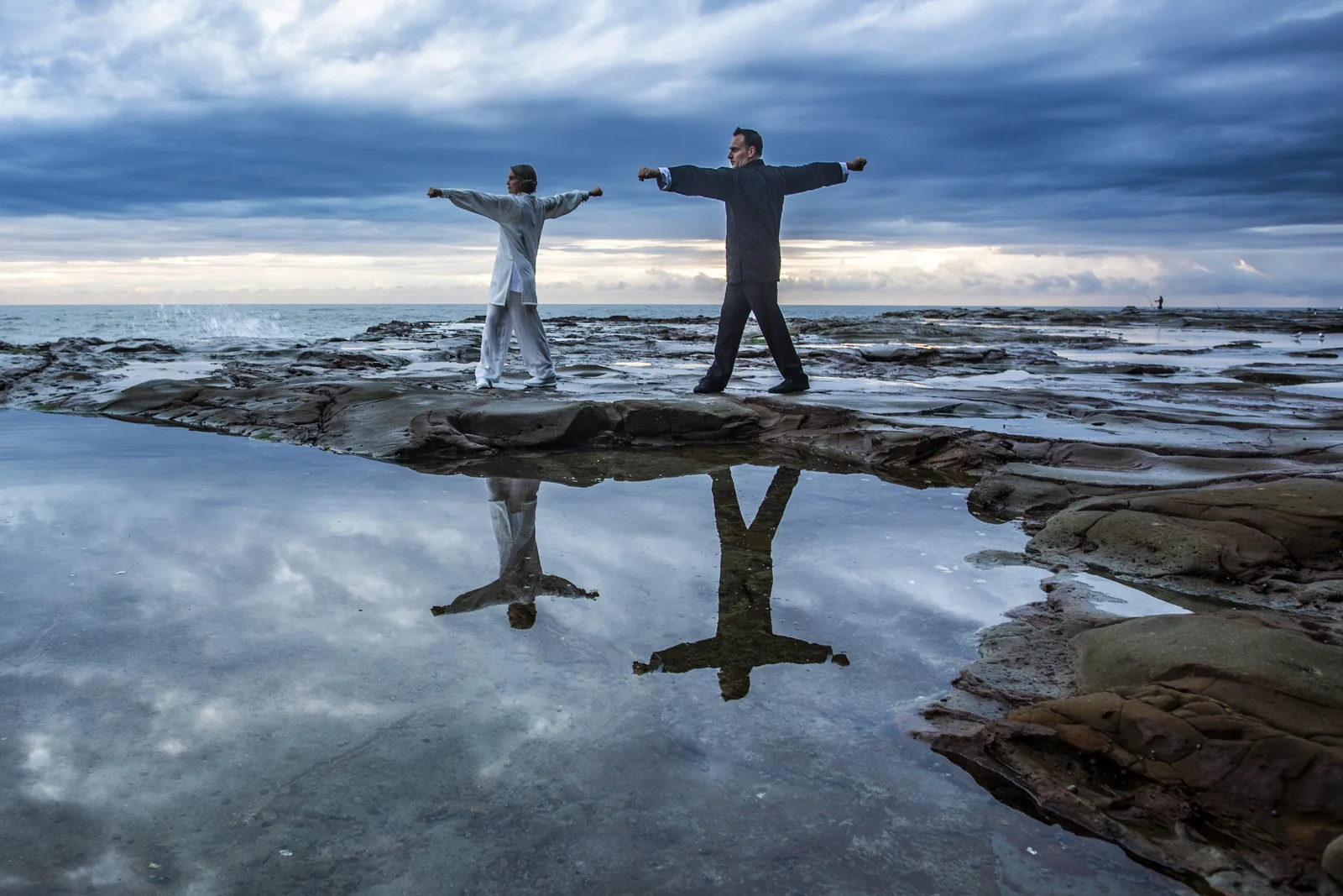 Two Tai Chi practitioners with arms outstretched on a sea rock platform at dawn.